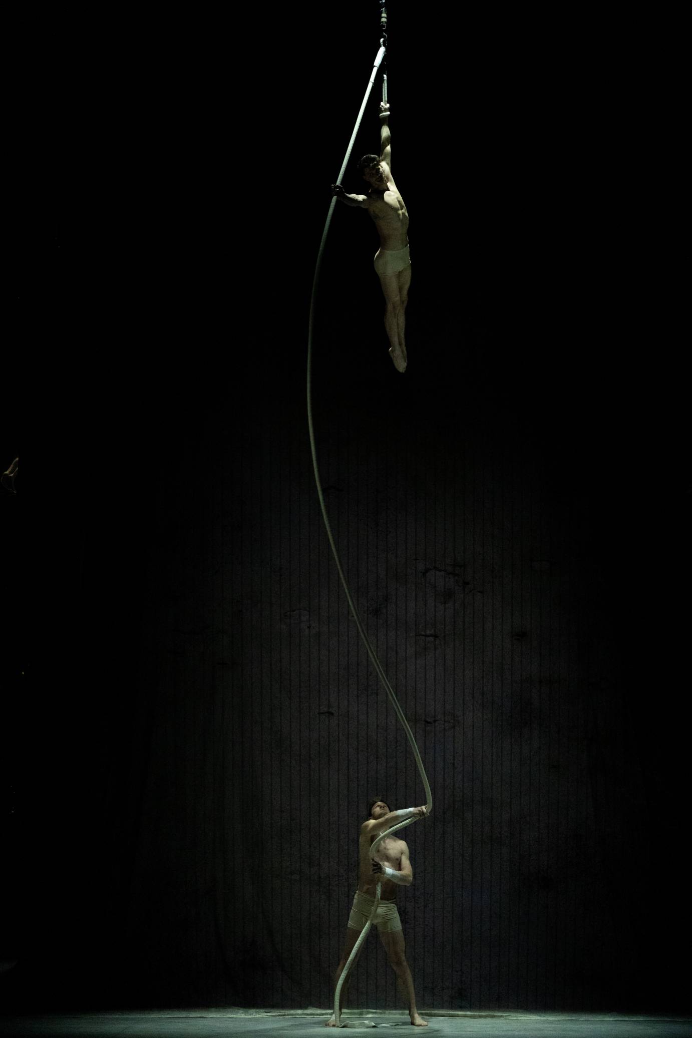 One man stands on a trapeze while another stands below him